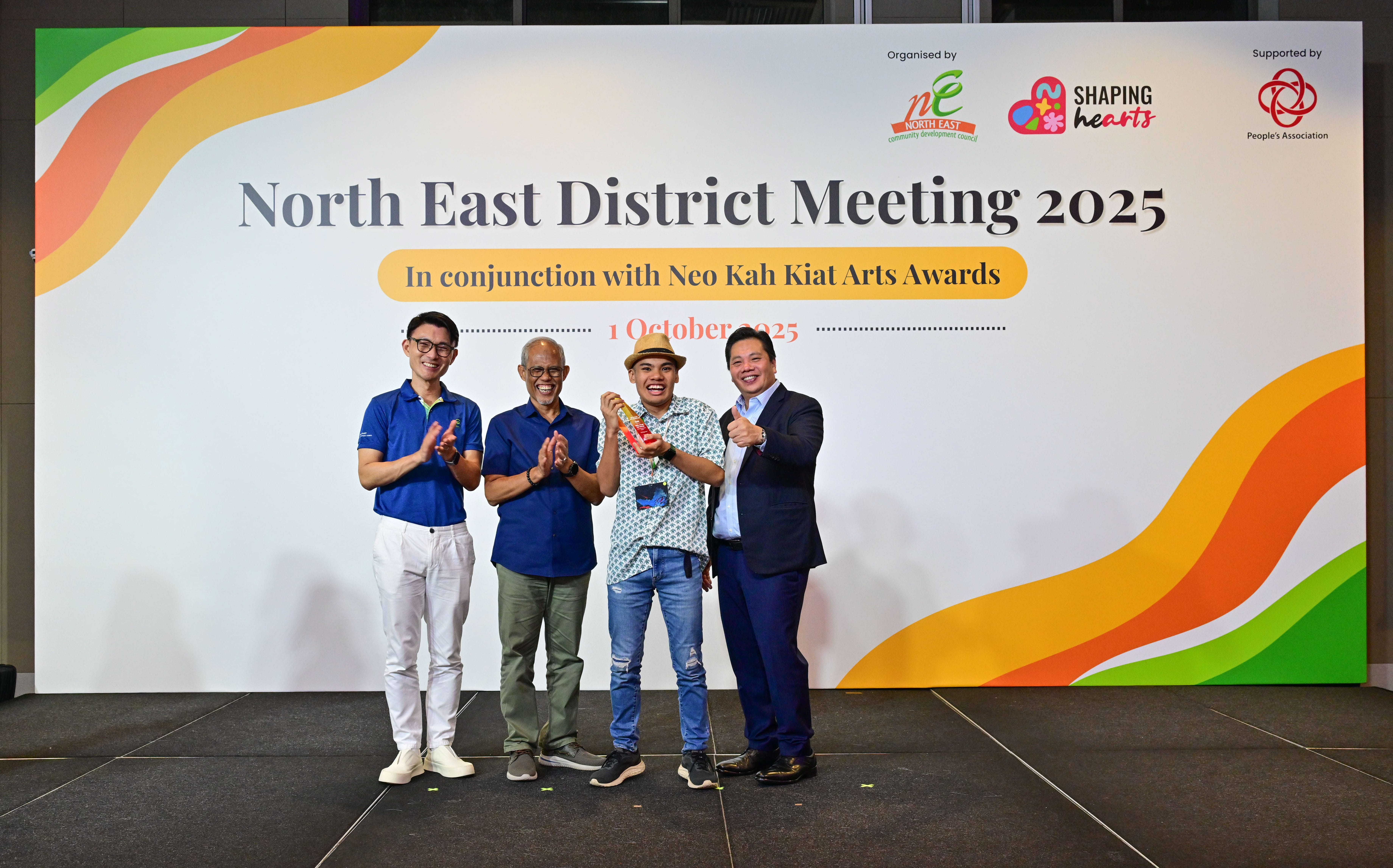 Award recipient standing on stage with the presenter, holding a acrylic plaque and posing for a group photo during the award ceremony, with a backdrop reading ‘North East District Meeting 2025 In conjunction with Neo Kah Kiat Arts Awards'
